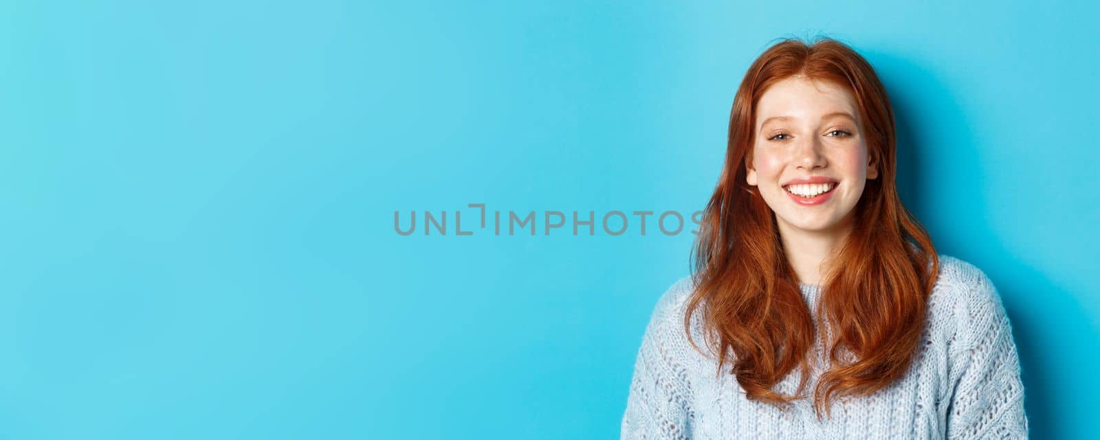 Close-up of cheerful teenage girl with red curly hair, smiling happy at camera, standing against blue background by Benzoix