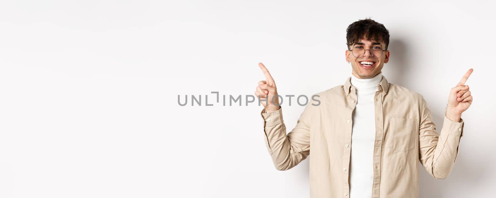 Image of smiling handsome man in glasses pointing fingers sideways, showing advertisements or variants, standing cheerful on white background by Benzoix