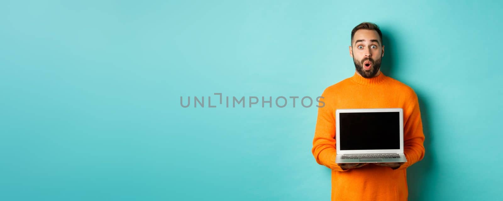 Handsome bearded man in orange sweater showing laptop screen, demonstrating promo, standing over light blue background.