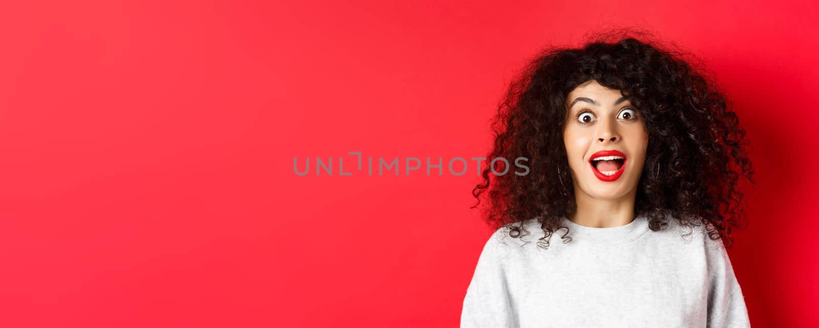 Close-up portrait of excited woman with curly hair, scream surprised and amazed, checking out special deal, standing on red background.
