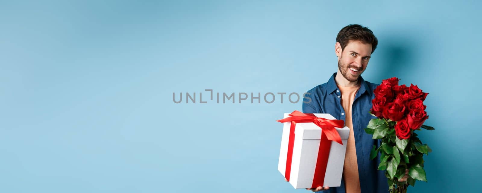 Love and Valentines day concept. Romantic smiling man giving you gift box and bouquet of flowers on date, standing over blue background by Benzoix