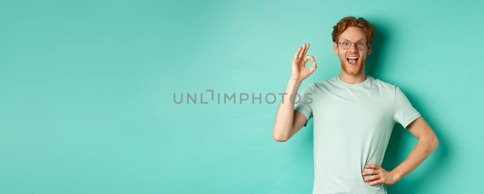 Amused young man with red hair, wearing glasses and t-shirt, showing okay sign and smiling excited, checking out something and approving it, standing over turquoise background.