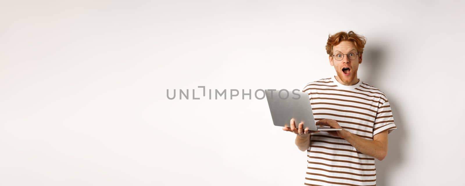 Impressed young man holding laptop in hands, staring at camera with excited face, reading online promotion on website, standing against white backround.