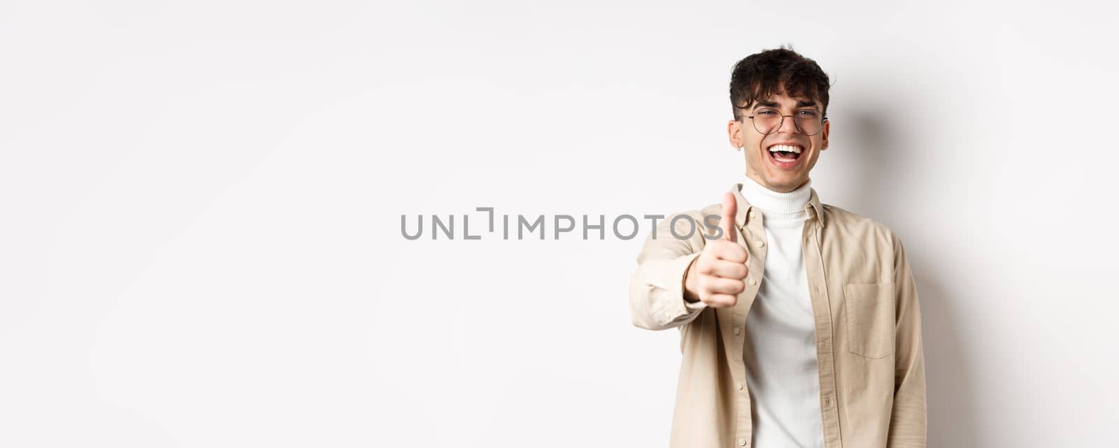 Nice one. Happy young man laughing and showing thumb up, like something good, standing on white background, complimenting you.