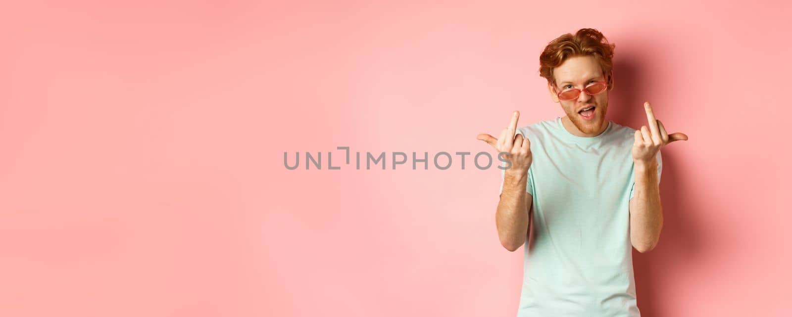 Arrogant and careless redhead man in sunglasses dont give a fuck, showing middle fingers at camera and frowning, standing over pink background by Benzoix