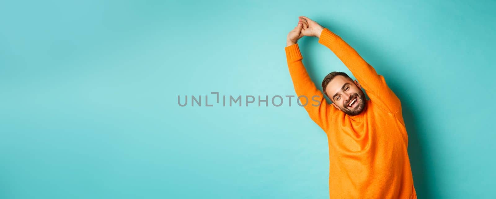 Image of handsome young man stretching hands and smiling after good rest, standing in orange sweater over light blue background.