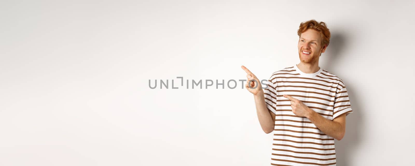 Cheerful caucasian guy with red hair and beard, looking and pointing left at logo banner, standing over white background by Benzoix