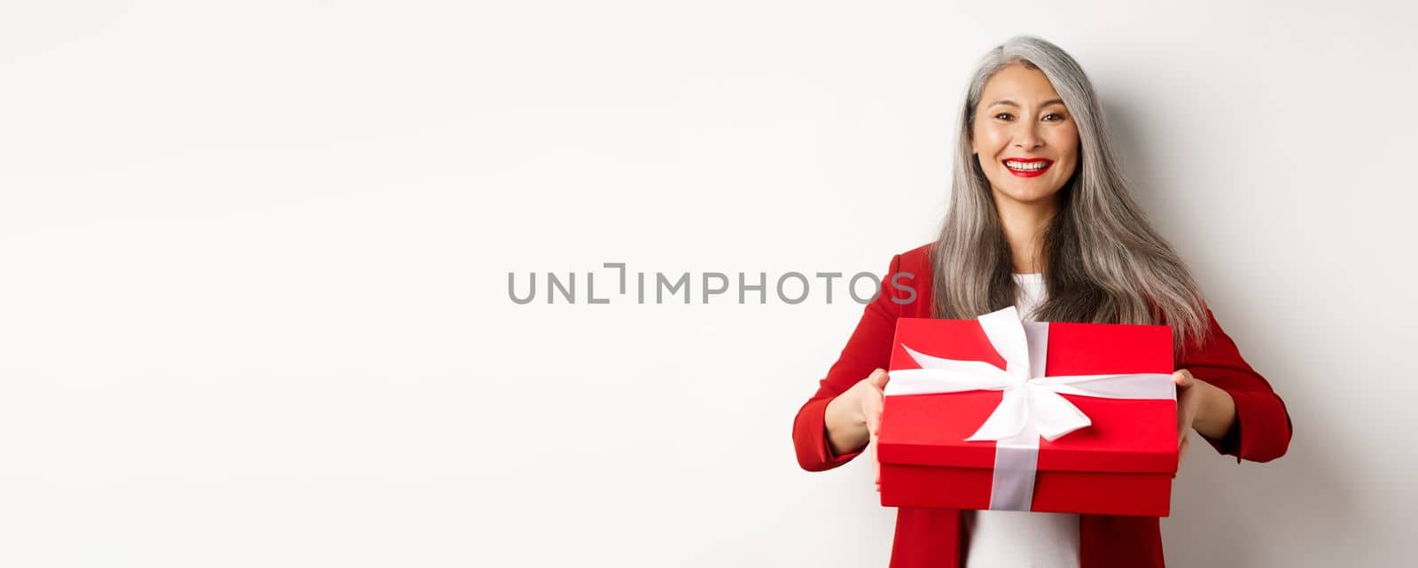 Elegant senior woman giving you present. Asian lady holding red gift box and smiling, standing over white background.