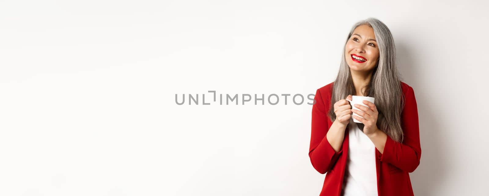 Business people concept. Smiling asian businesswoman having coffee break, holding warm mug and looking upper left corner dreamy, white background.