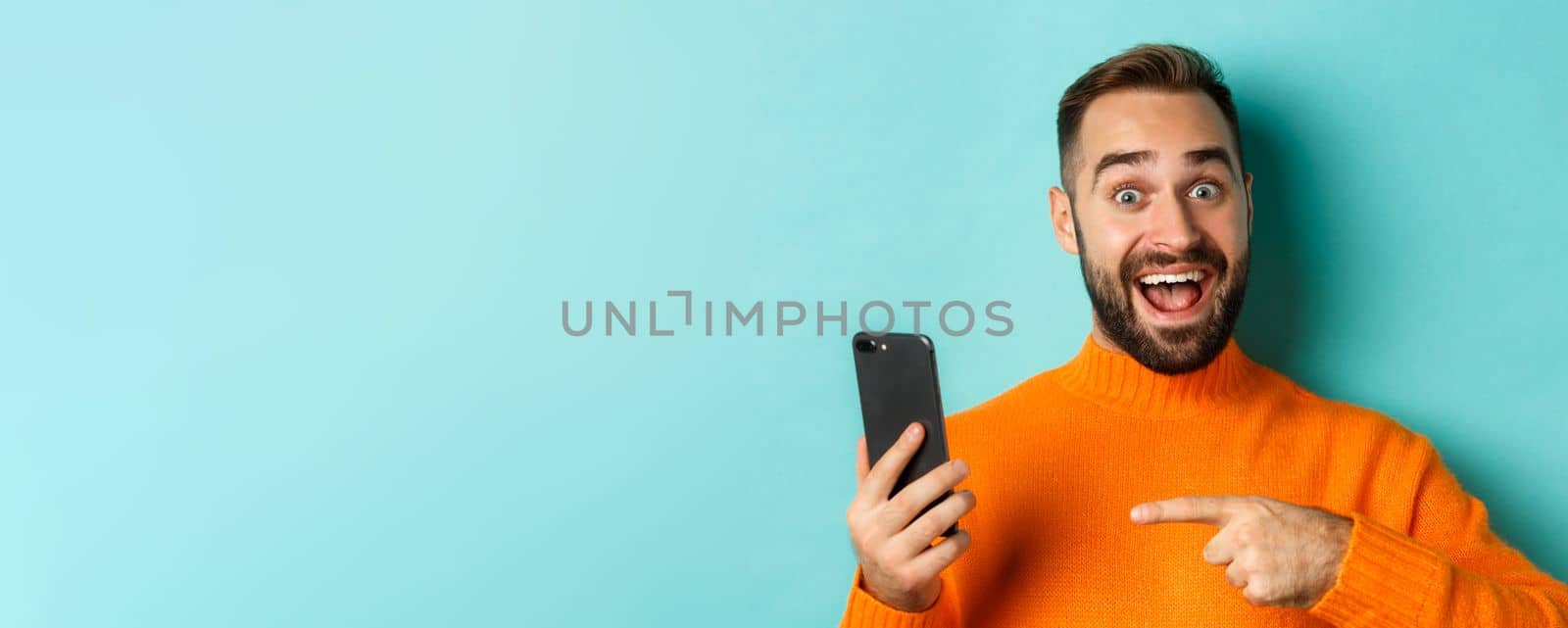 Close-up of impressed man showing something on mobile phone, pointing at smartphone and gasping amazed, standing in orange sweater over light blue background by Benzoix