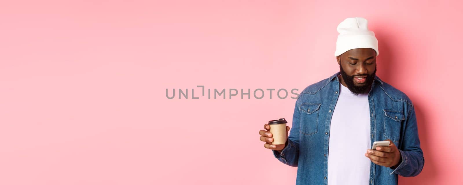 Image of stylish Black man hipster drinking takeaway coffee, reading message on phone, standing over pink background.
