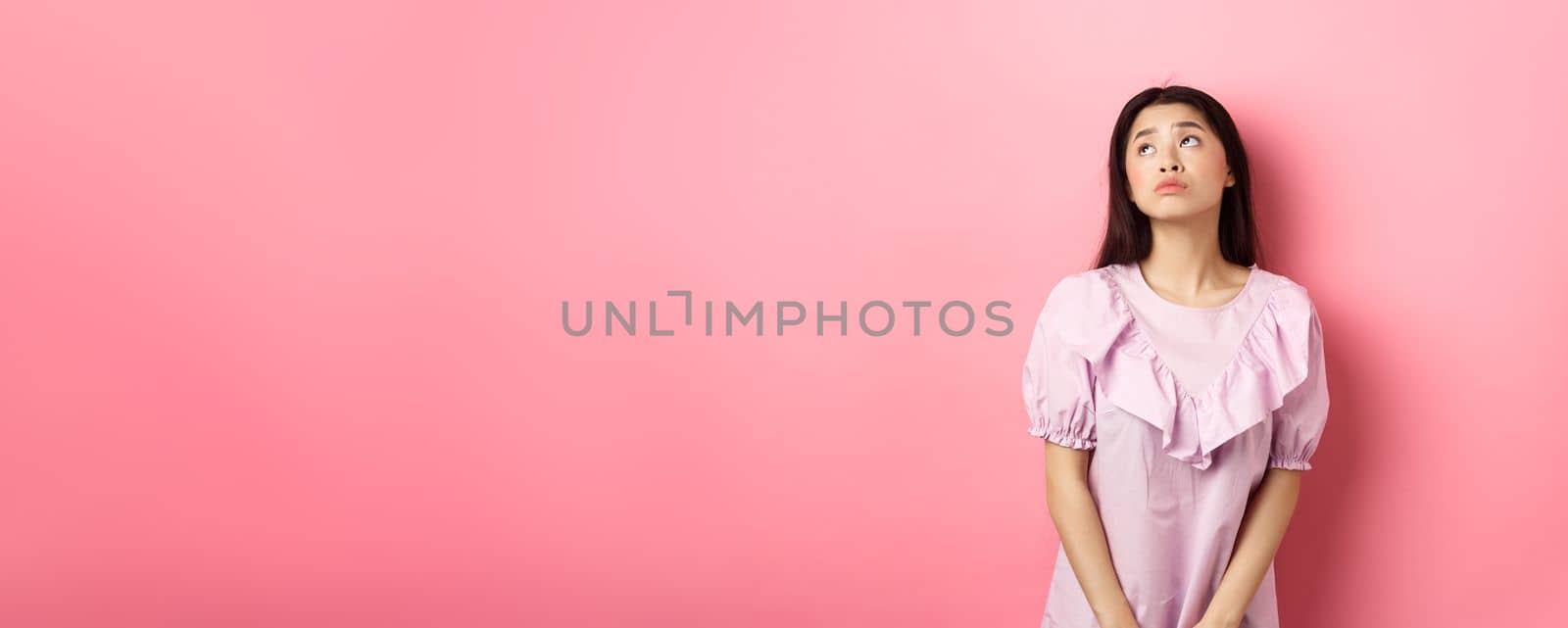 Dreamy and sad asian woman look at upper left corner logo, standing pensive and gloomy in pink romantic dress, white background by Benzoix