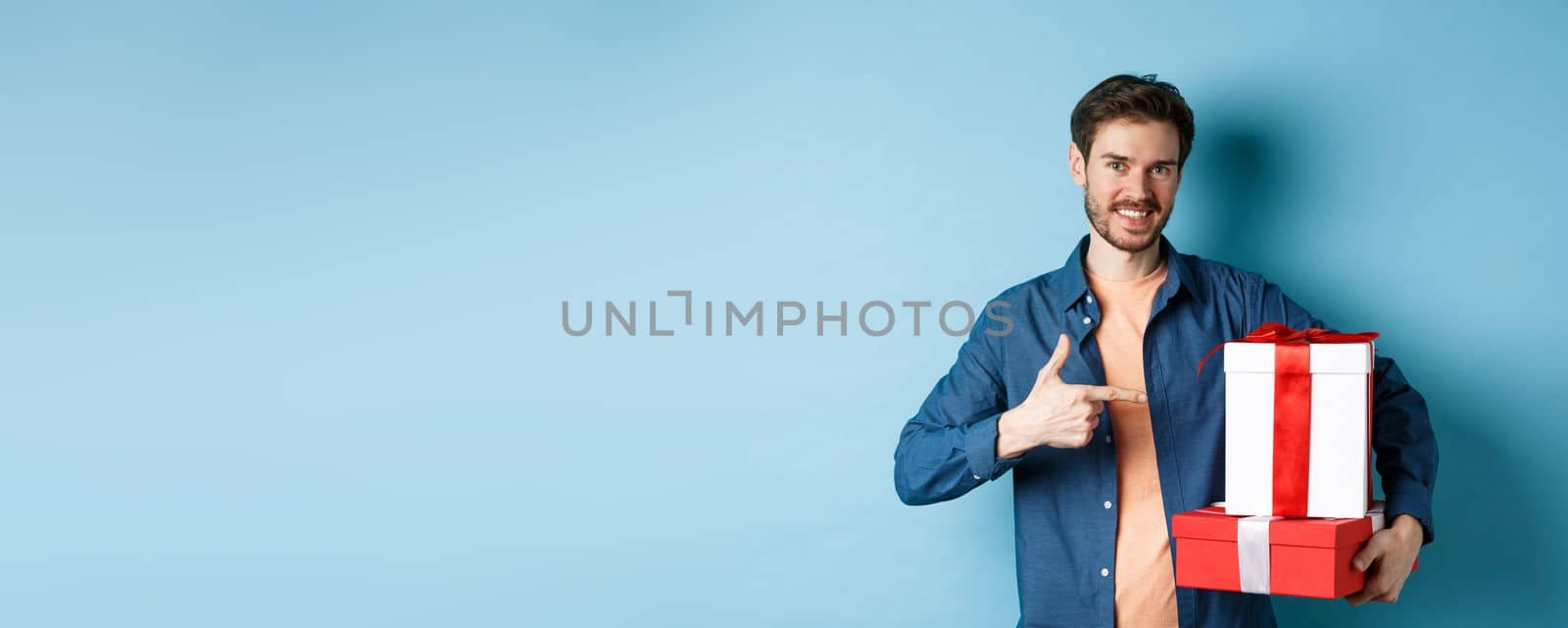 Attractive boyfriend pointing finger at valentines day surprise gifts, showing romantic presents for girlfriend, standing against blue background.