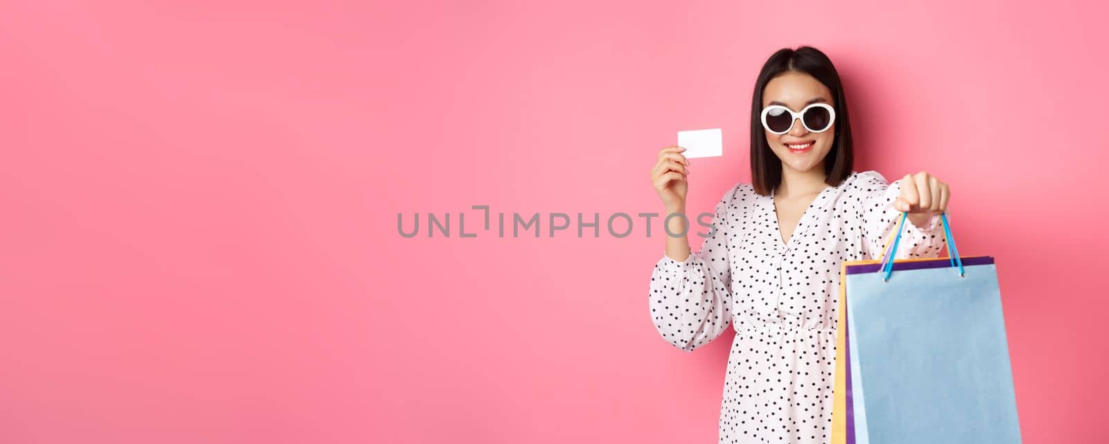 Beautiful asian woman in sunglasses going shopping, holding bags and showing credit card, standing over pink background.