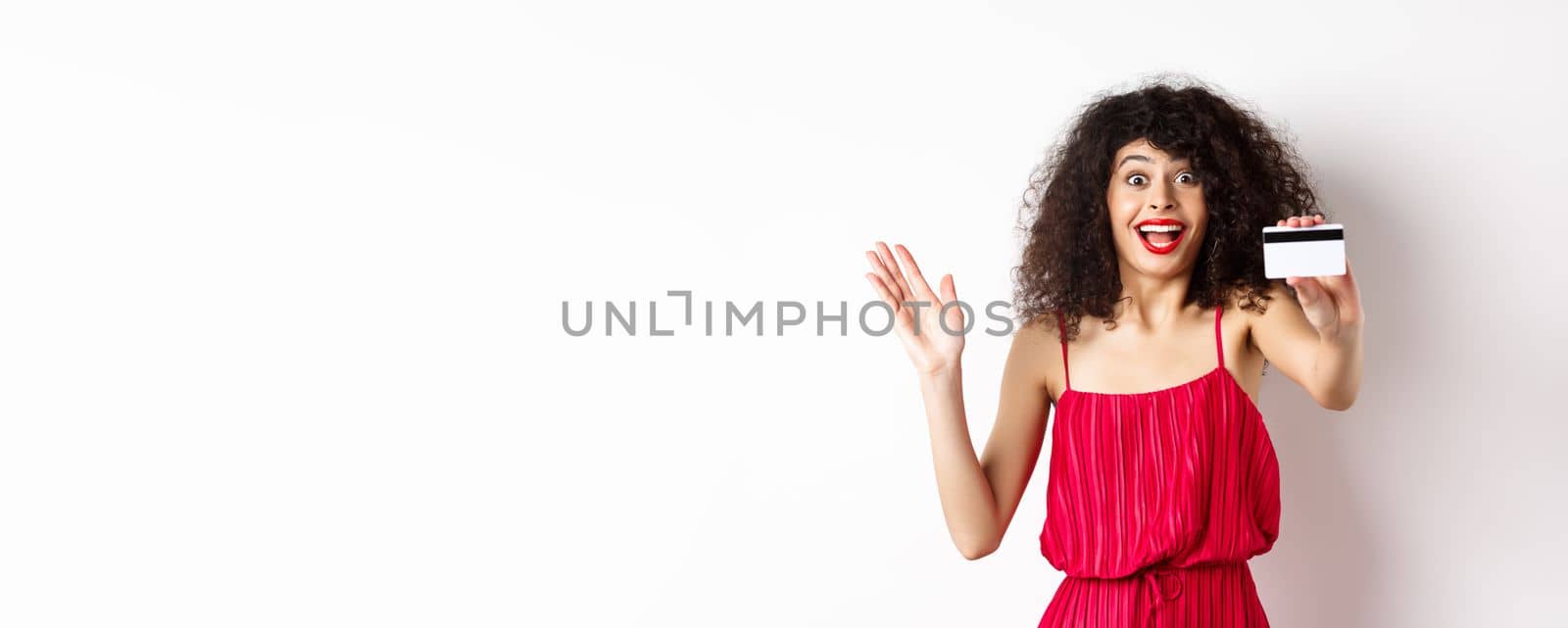 Shopping. Amazed curly-haired woman in red dress, showing plastic credit card and scream from excitement, standing on white background by Benzoix