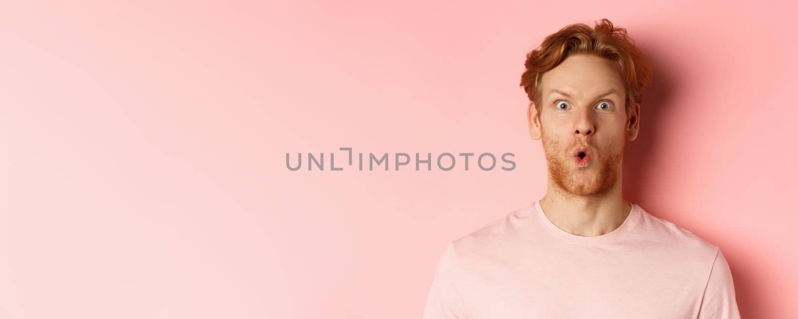 Headshot of young redhead man with beard, saying wow and staring at camera amazed, checking out promo deal, standing over pink background by Benzoix