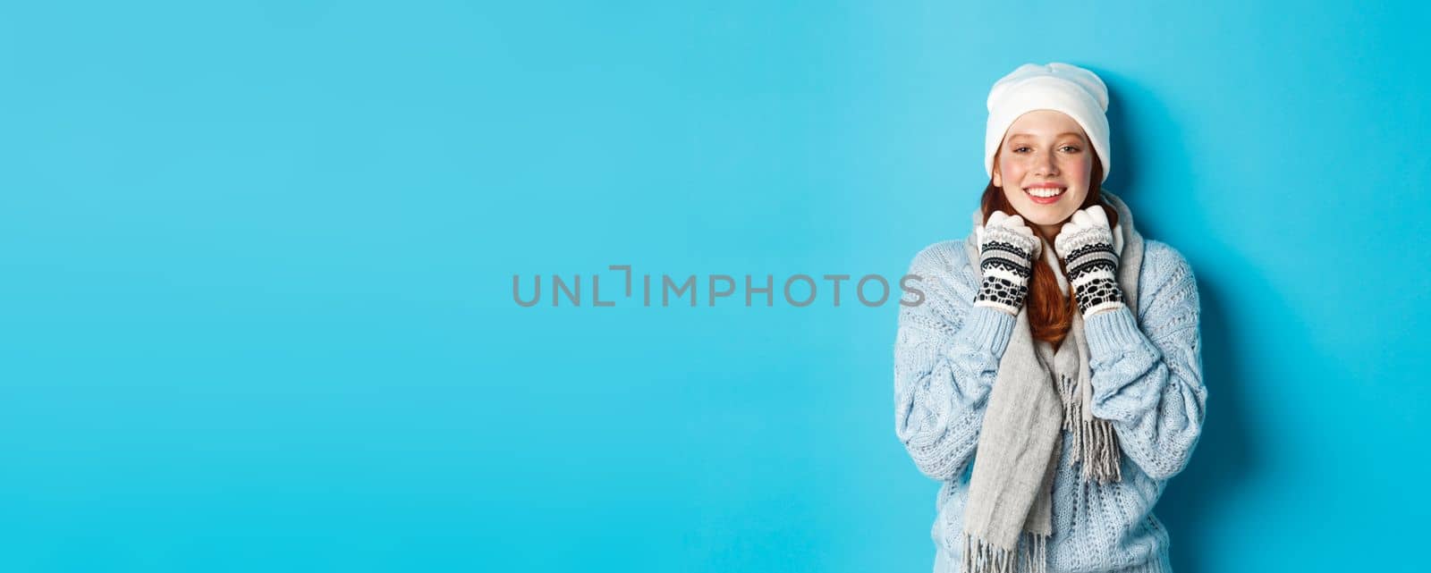 Winter and holidays concept. Cute redhead girl in white beanie and gloves smiling at camera, looking delighted, standing against blue background.