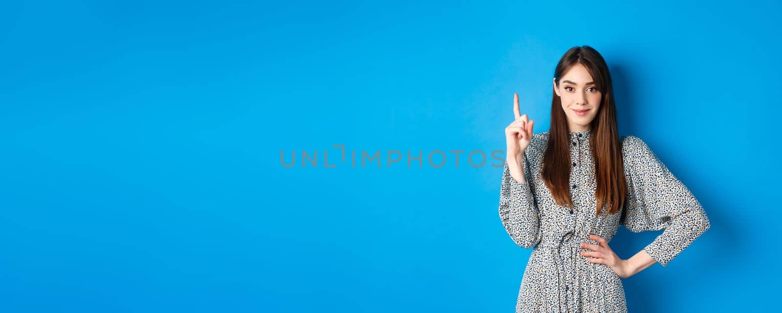 Young beautiful lady in dress with natural long hair, showing number one with finger and smiling, standing against blue background by Benzoix