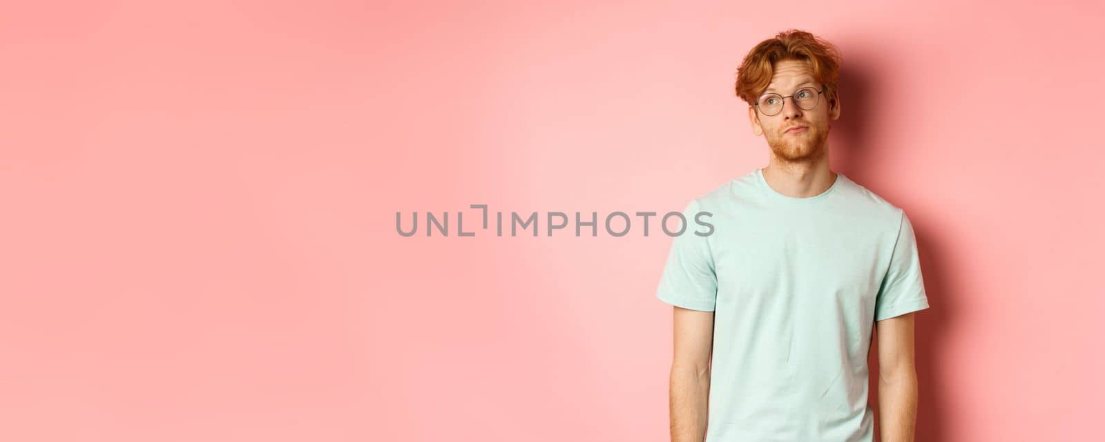 Image of young redhead man with beard, wearing t-shirt and glasses, looking left with bored unbothered face, standing over pink background.