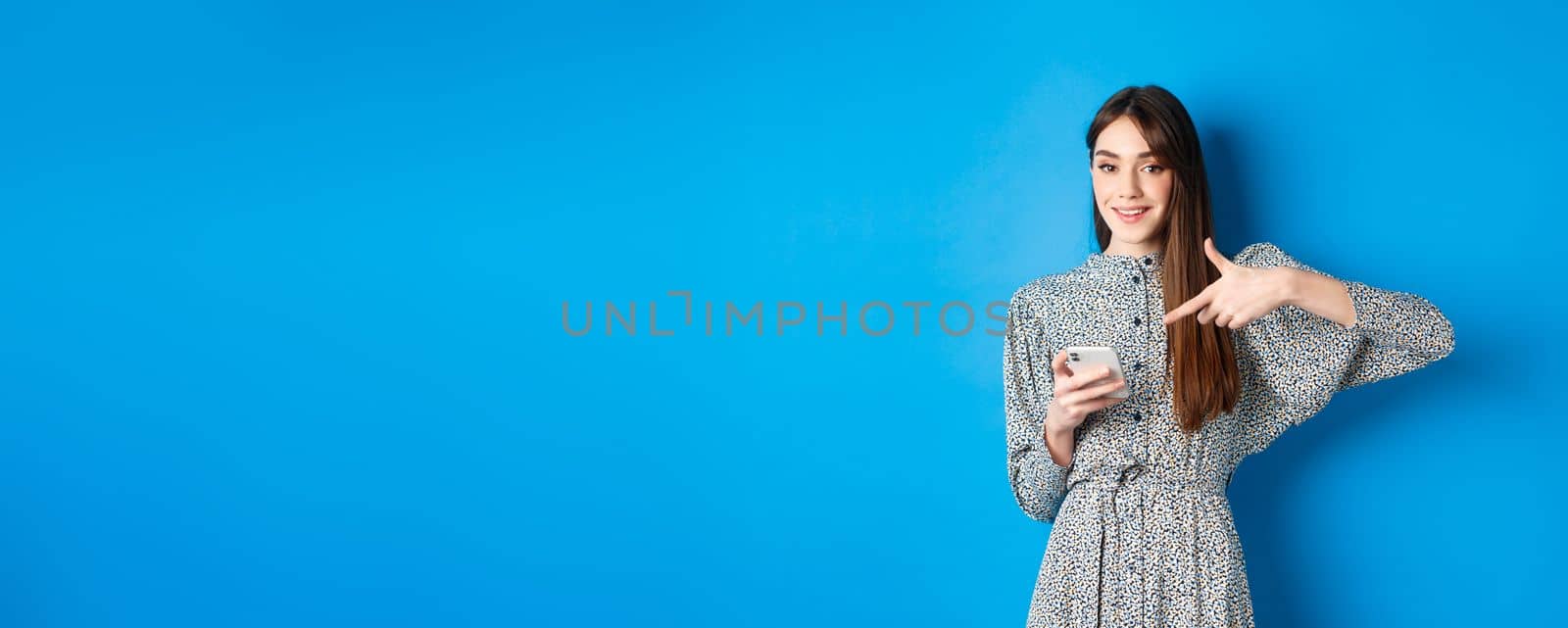 Stylish caucasian woman in dress, pointing at smartphone and smiling, showing online promo, standing on blue background.