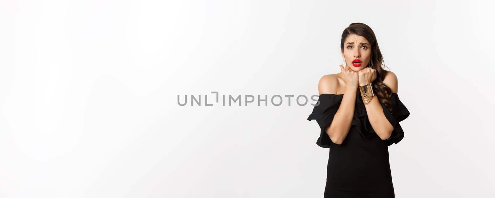 Fashion and beauty concept. Scared timid woman grimacing, looking concerned and worried at camera, standing over white background in black dress.