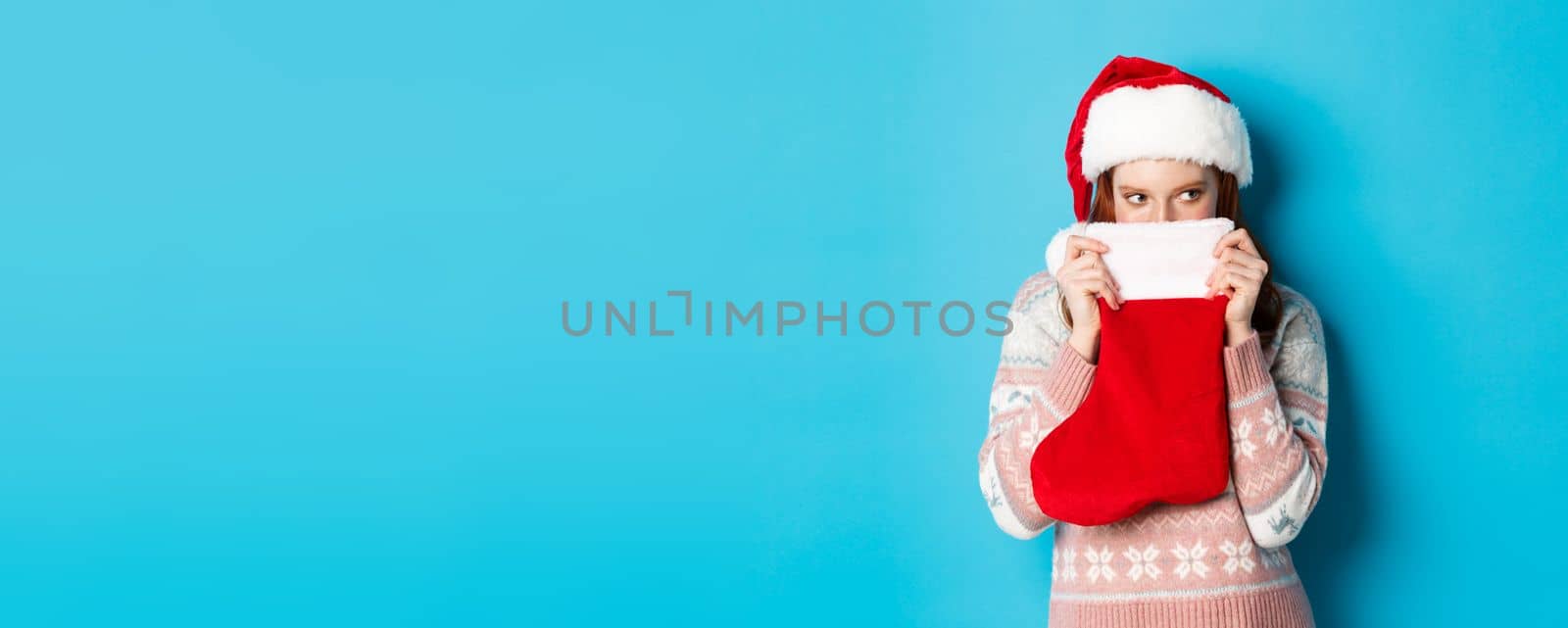 Cute girl cover face with christmas stocking, staring right with cunning gaze, standing in Santa hat and celebrating winter holidays, blue background by Benzoix
