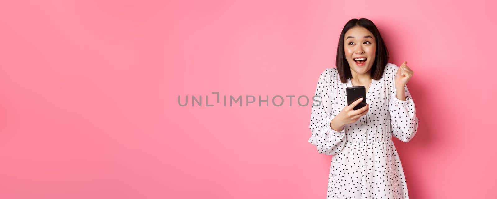 Online shopping and beauty concept. Excited asian woman winning in internet, holding smartphone and rejoicing, smiling happy and celebrating, standing over pink background.