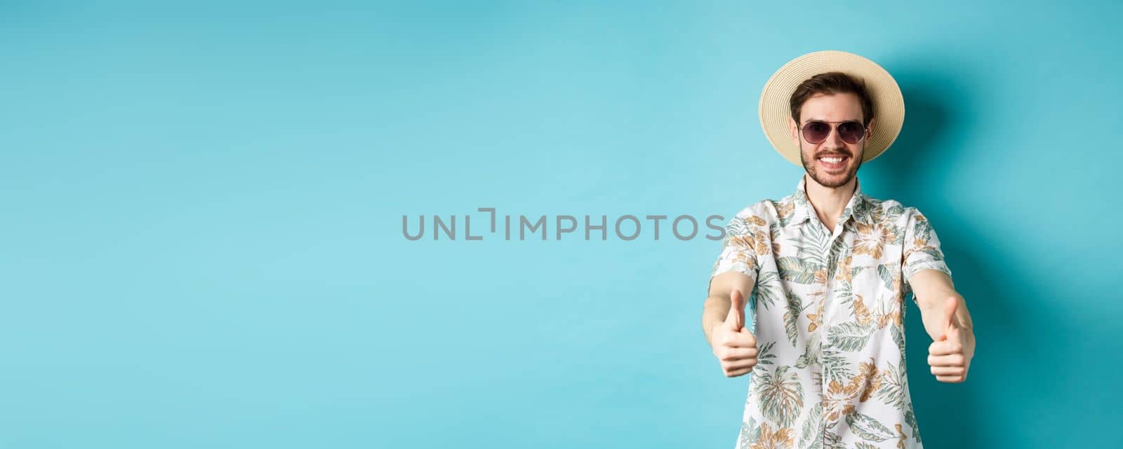 Happy tourist in sunglasses and straw hat showing thumbs up and smiling, enjoying summer holiday, standing on white background by Benzoix