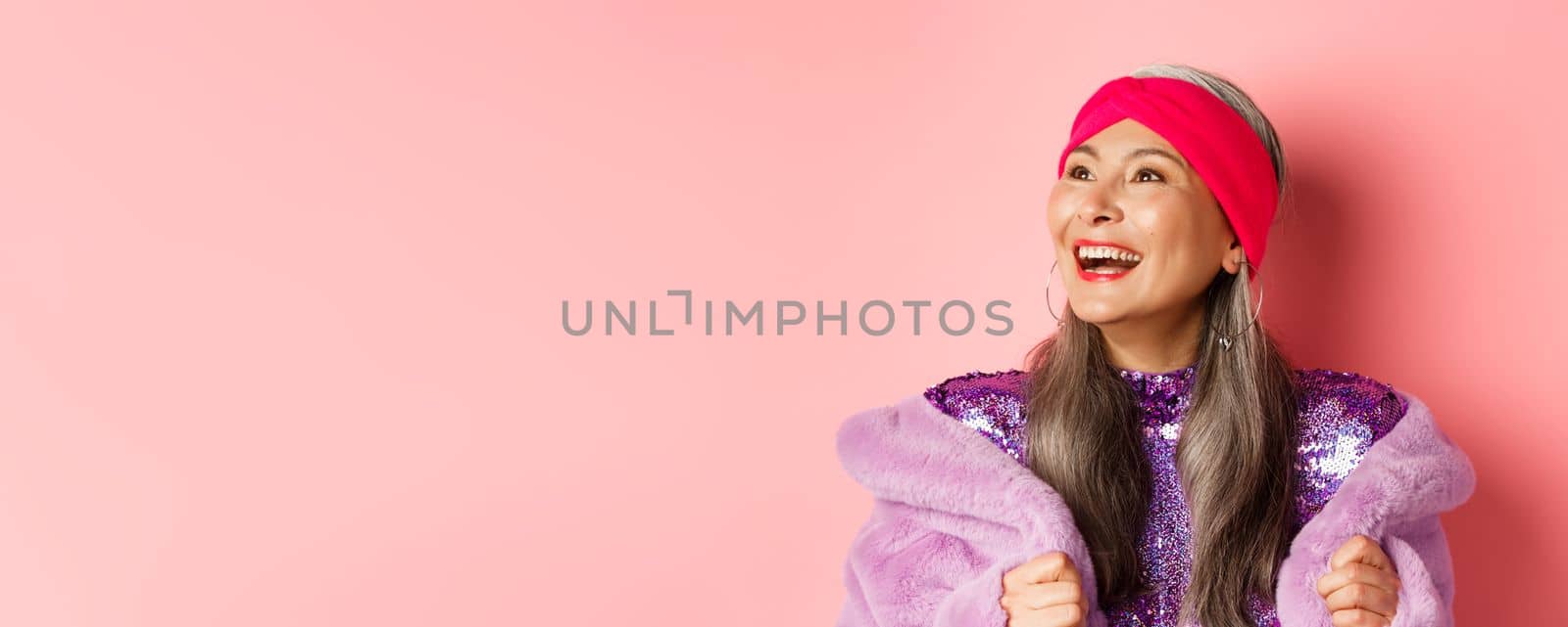 Fashion. Close-up of happy asian senior woman smiling, looking left with cheerful face, standing in purple faux fur coat, pink background by Benzoix