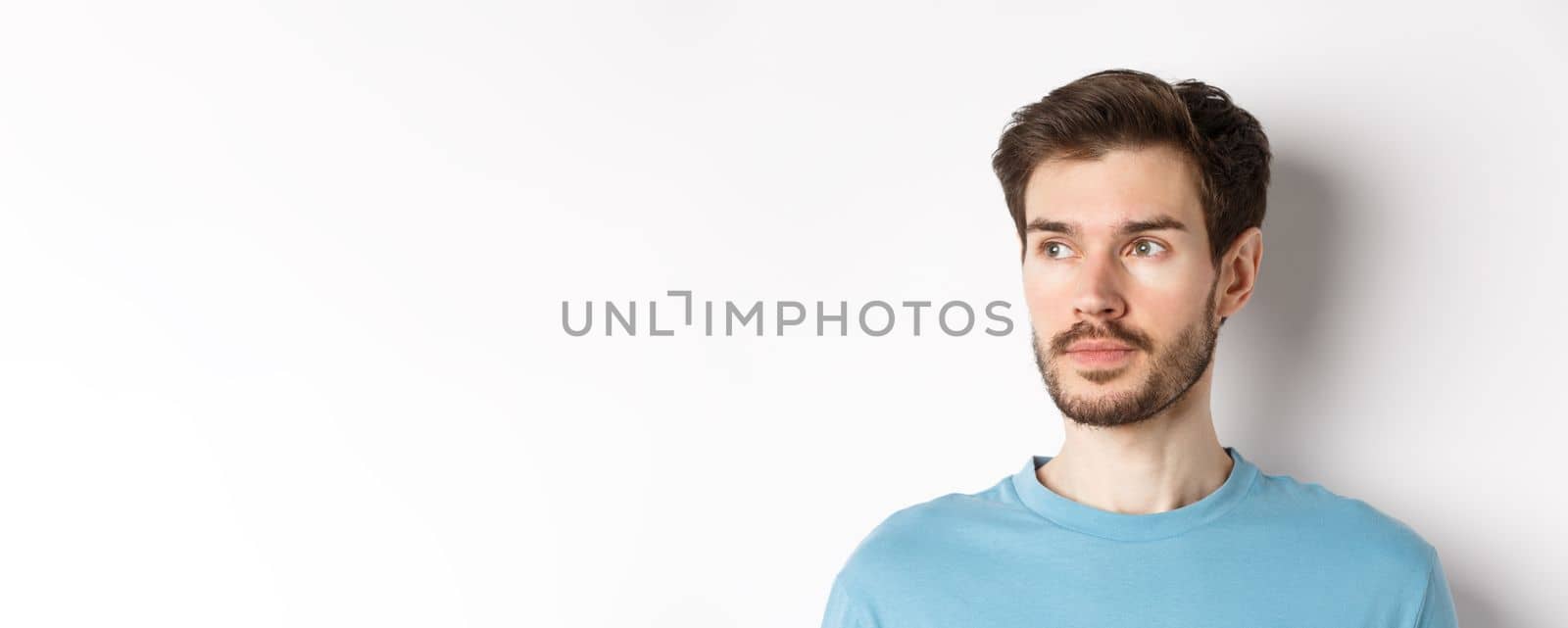 Close-up of handsome male model with beard and moustache, looking left with thoughtful face, standing over white background by Benzoix
