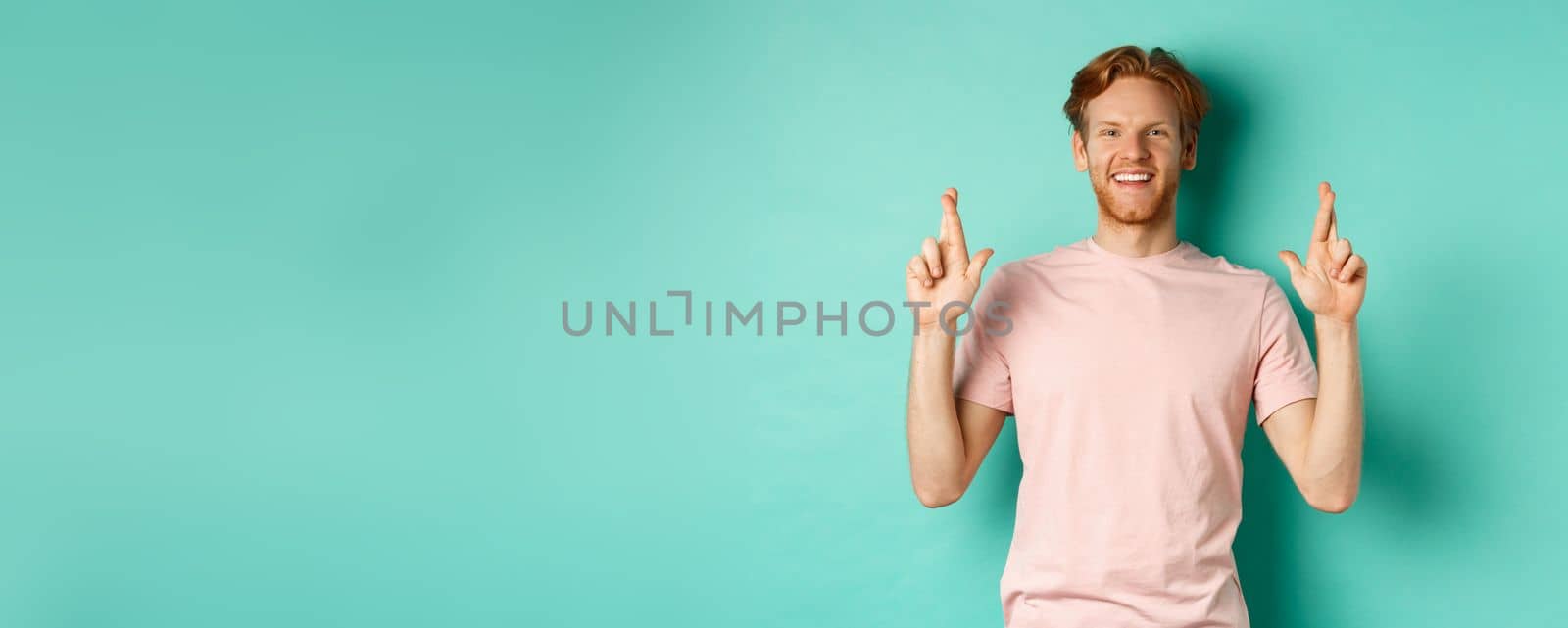 Optimistic guy with red hair and beard smiling, cross fingers for good luck and looking hopeful at camera, making a wish, standing over mint background.