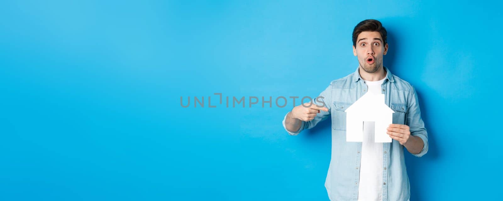 Insurance, mortgage and real estate concept. Surprised young man pointing at house card model and looking at camera, standing against blue background by Benzoix