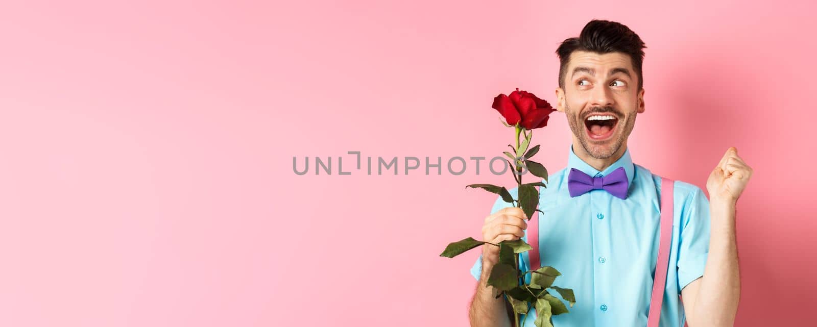 Romance and Valentines day concept. Cheerful man in bow-tie screaming from happiness, holding red rose and jumping on date, standing over pink background.