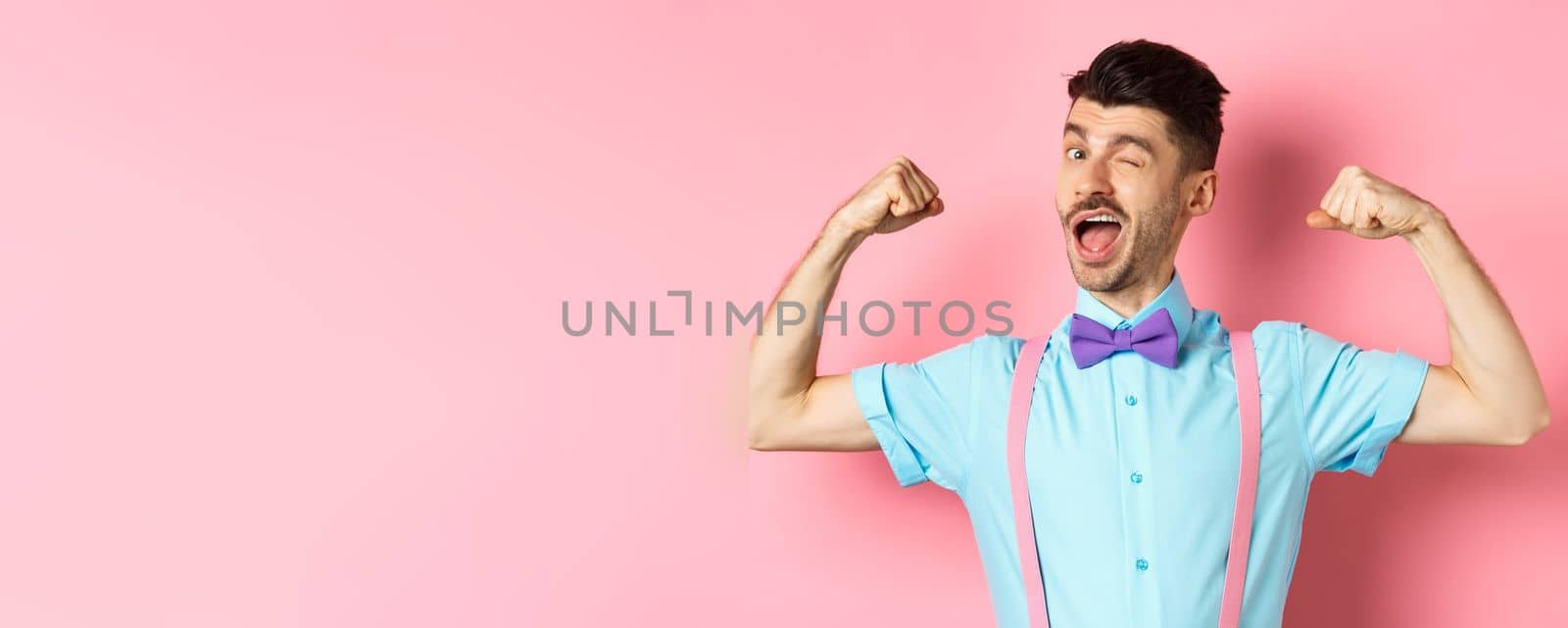 Strong and funny guy with french moustache, flexing biceps and winking at camera, show-off his strengths, standing over pink background.
