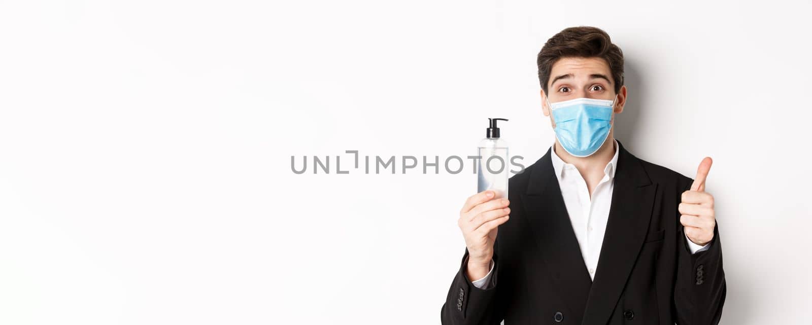 Concept of covid-19, business and social distancing. Close-up of satisfied handsome man in suit and medical mask, showing thumb-up and hand sanitizer, standing against white background by Benzoix