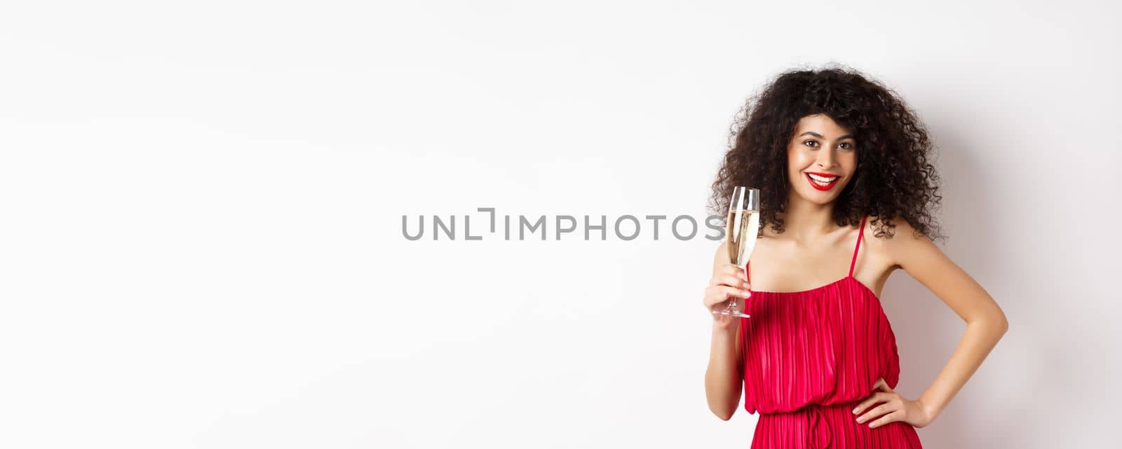 Sassy young woman in elegant red dress, posing with glass of champagne on white background, having romantic date on valentines day.