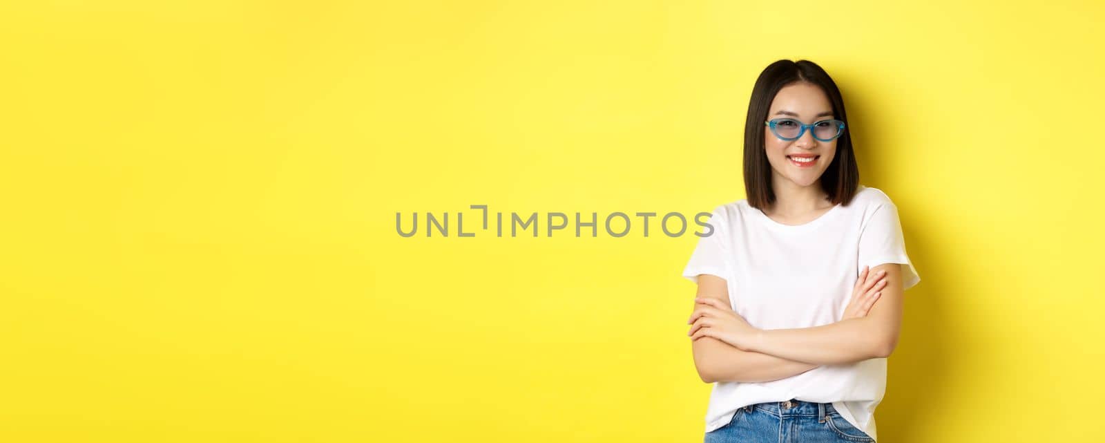 Fashion and lifestyle concept. Sassy and confident asian woman in trendy sunglasses looking self-assured at camera, standing over yellow background by Benzoix