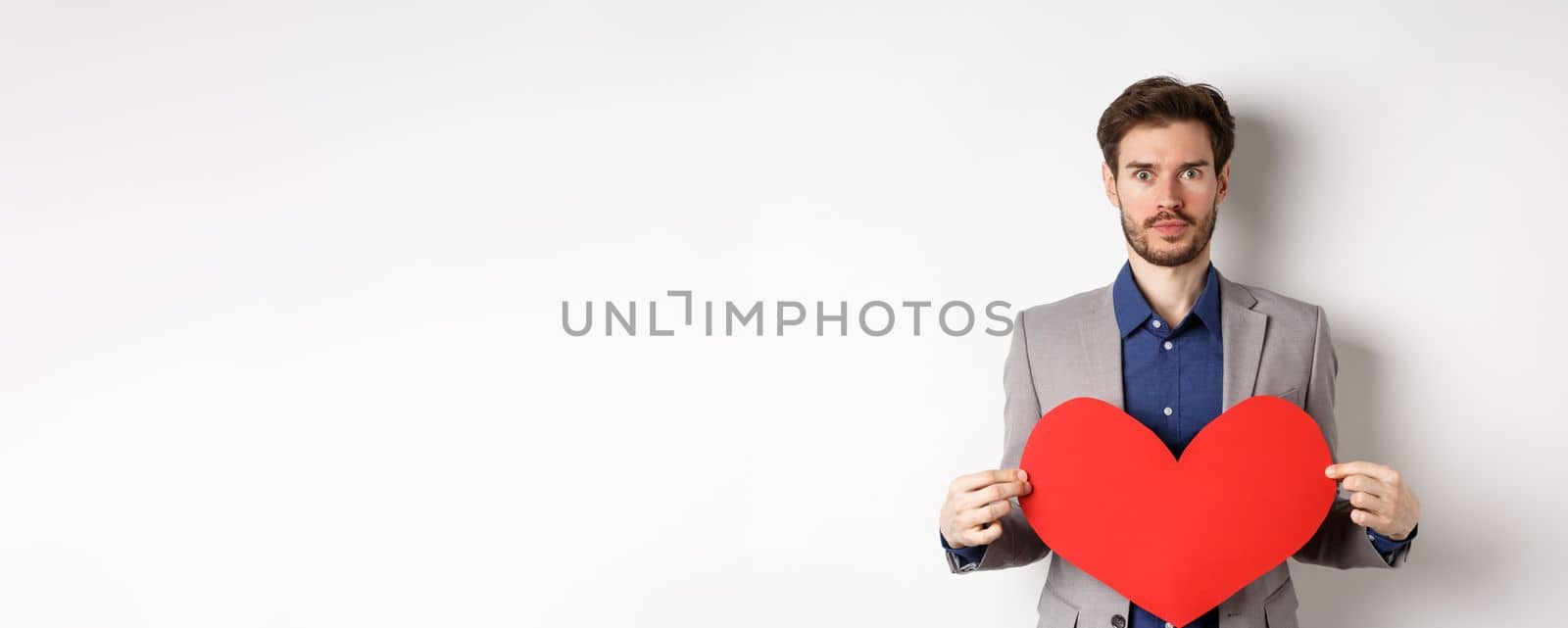 Excited caucasian man in suit looking at camera, holding big red heart cutout on valentines day, standing over white background by Benzoix