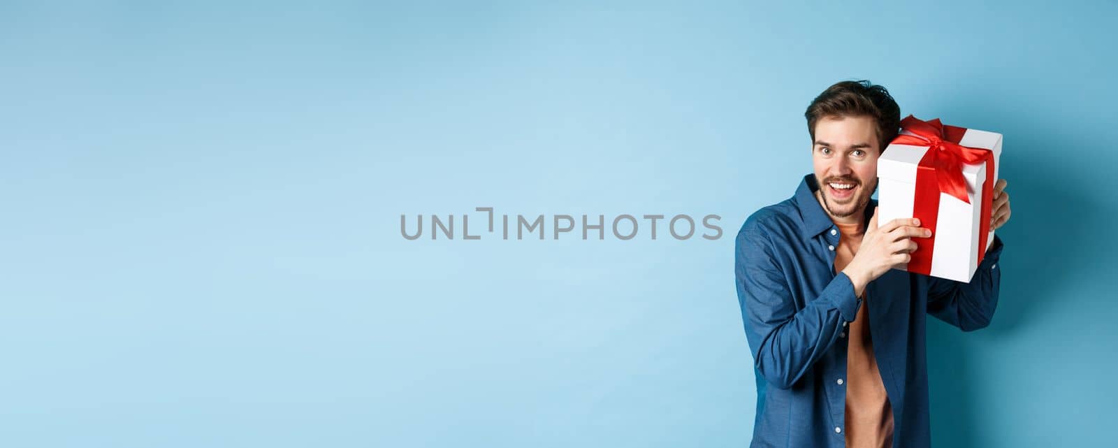 Valentines day. Happy young man got present on special holiday, trying guess what inside gift box and smiling, standing over blue background by Benzoix