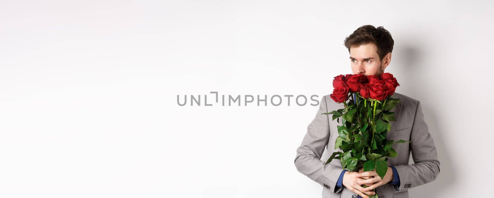 Romantic man in suit smell bouquet of roses and looking pensive, standing over white background. Concept of valentines day.