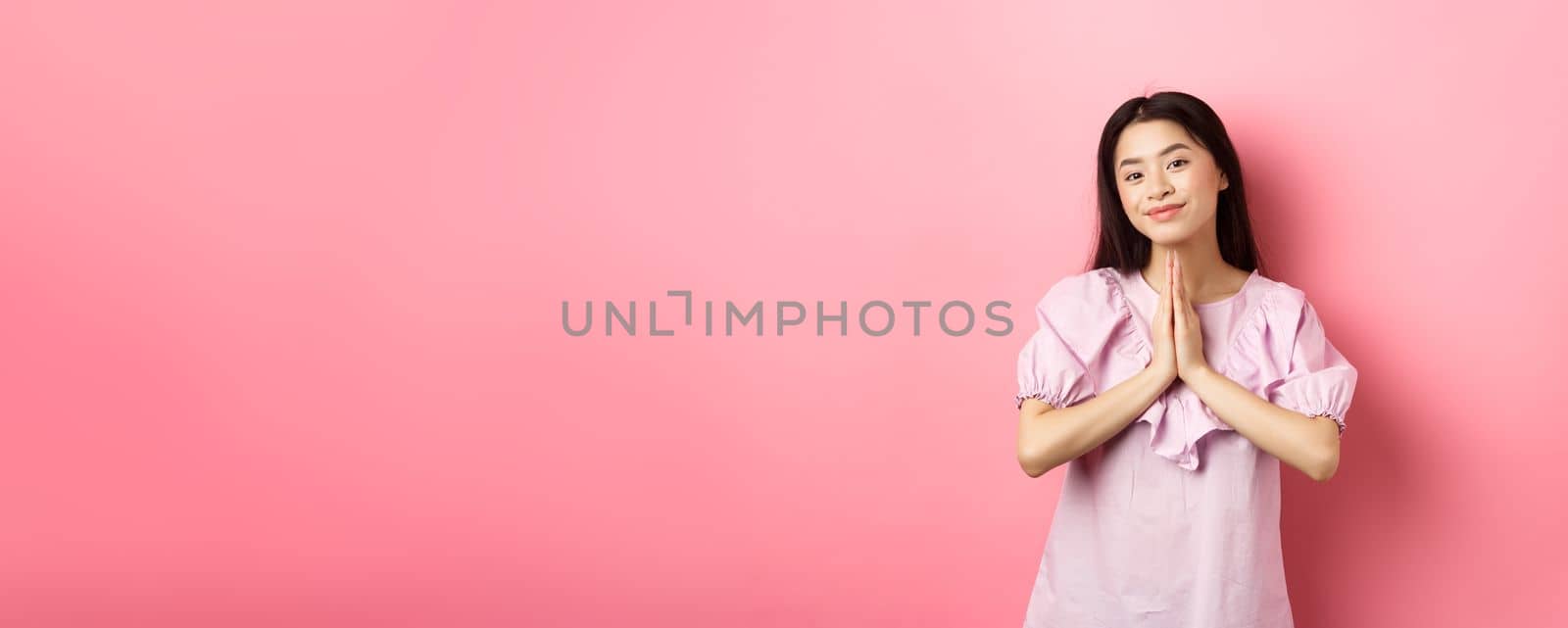 Cute asian girl say thank you, smiling and looking happy, showing namaste gesture in gratitude, standing in dress against pink background.