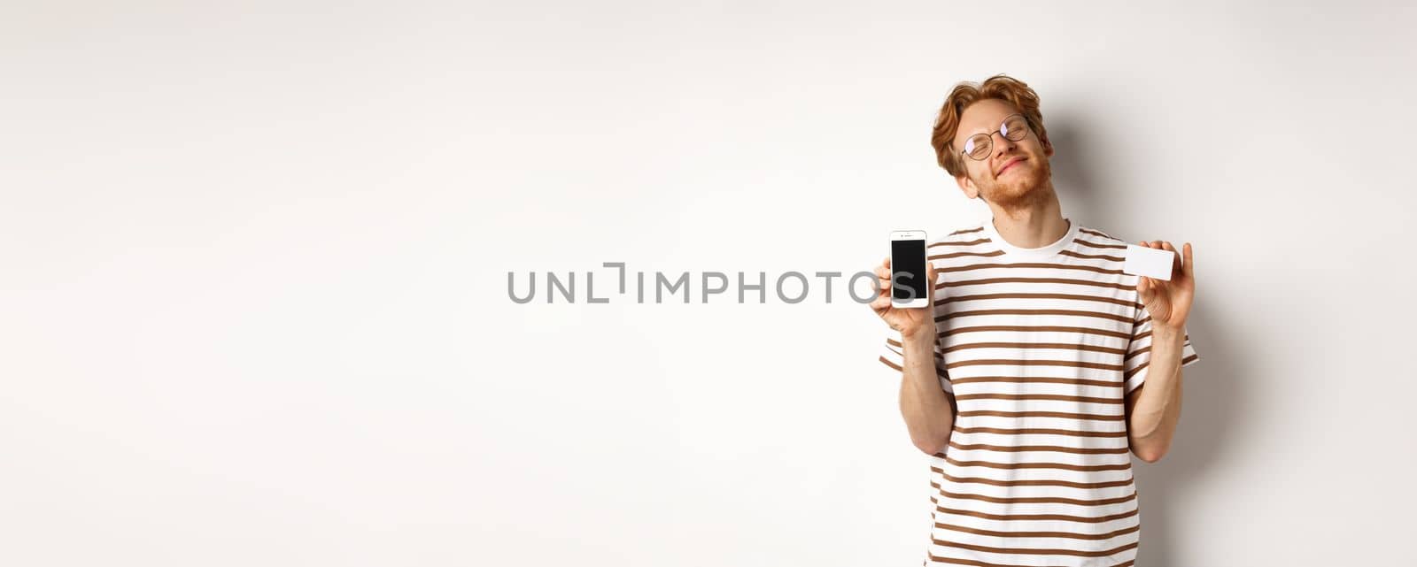 Shopping and finance concept. Pleased young man with red hair smiling from satisfaction, showing smartphone blank screen and credit card, white background.