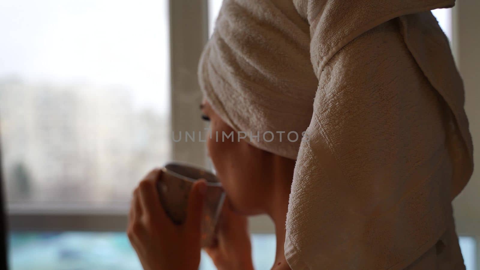 Middle aged woman looks good with bare shoulders in a white towel on her head holds a cup and drinks coffee or tea against the window.