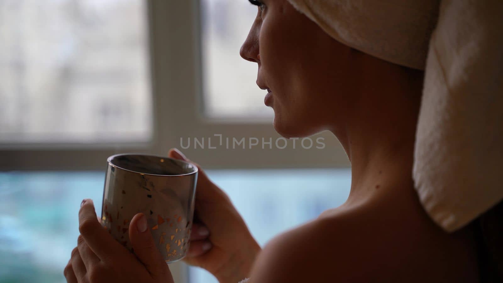 Middle aged woman looks good with bare shoulders in a white towel on her head holds a cup and drinks coffee or tea against the window by Matiunina