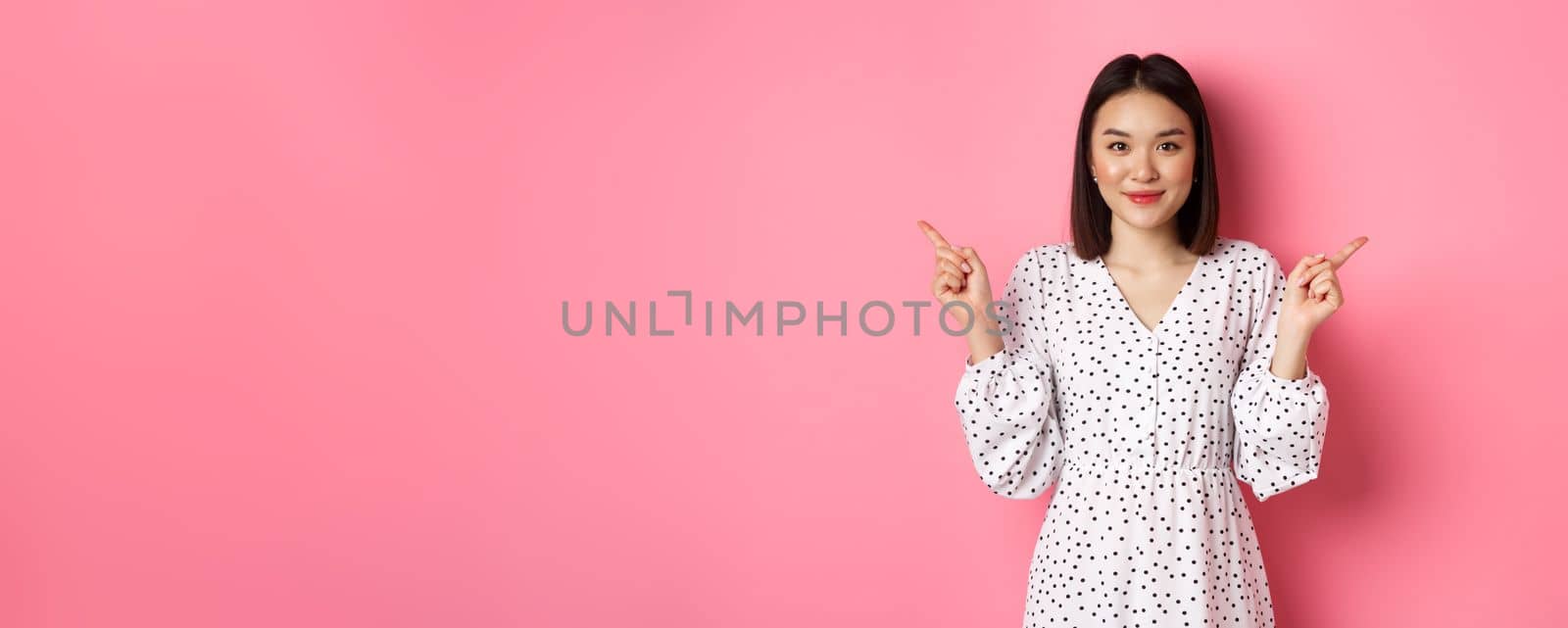 Beautiful asian woman making choice on shopping, pointing fingers sideways and showing variants, smiling at camera, standing over pink background by Benzoix