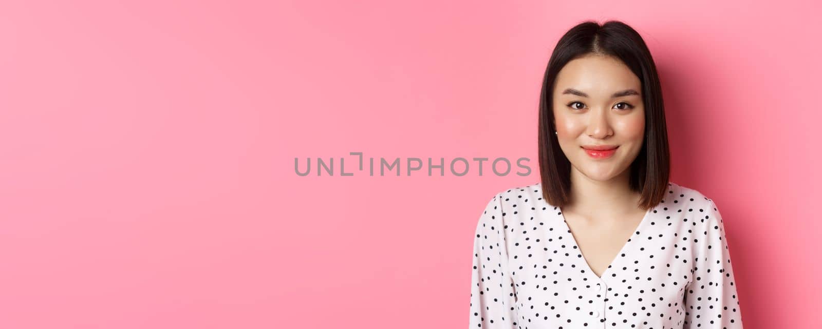 Close-up of cute asian female model looking at camera, blushing and smiling kawaii, standing over pink background.