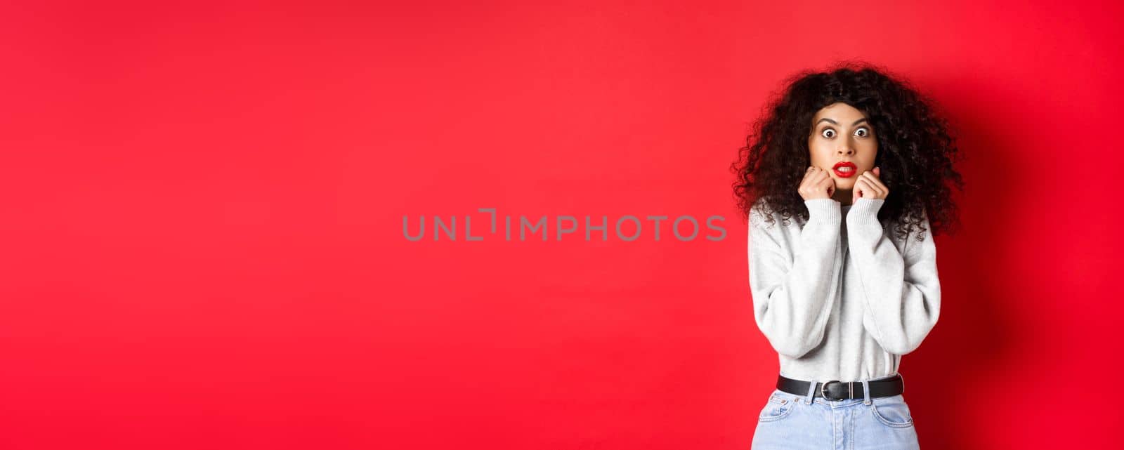 Shocked caucasian woman with curly hairstyle, looking at something impressive, gasping amazed, checking out promo offer, red background.