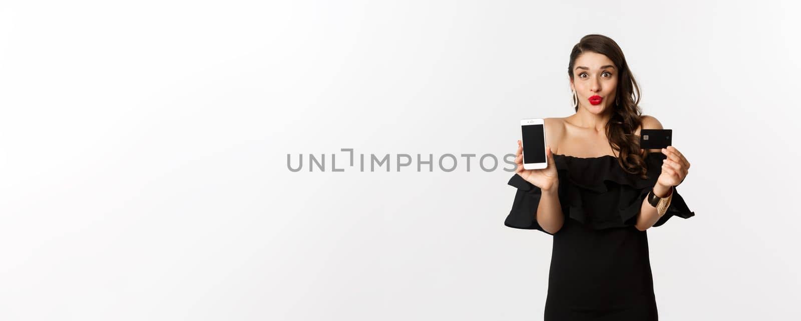 Fashion and online shopping concept. Happy young woman in black dress, showing credit card and mobile screen, standing over white background by Benzoix