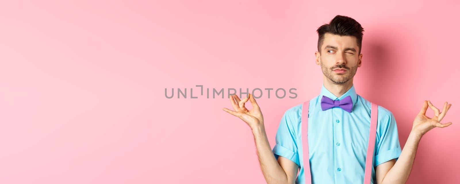Funny guy with moustache and bow-tie fake meditating, peeking aside while doing yoga asana, standing over pink background.