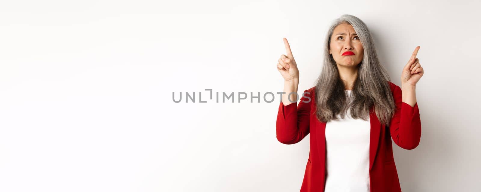 Disappointed asian senior woman looking, pointing fingers up and grimacing, dislike something, standing skeptical over white background.